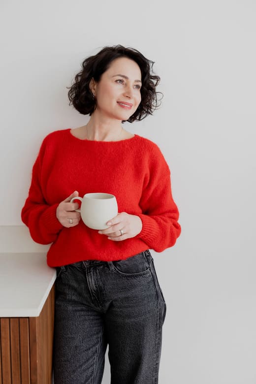 herzschaetzerin-claudia-di-chio-arbeitskultur-kennenlernen Herzschätzerin, Claudia di Chio mit rotem Pullover und weißem Becher in den Händen, der Blick abgewandt.