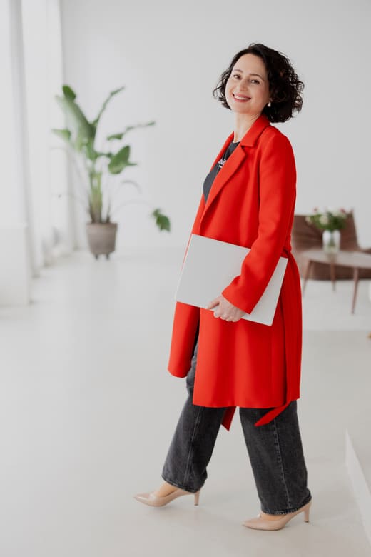herzschaetzerin-claudia-di-chio-gesunde-arbeitskultur-podcast Herzschätzerin, Claudia di Chio in rotem Mantel, Laptop unter dem Arm, seitlich gehend mit Blick zur Kamera.
