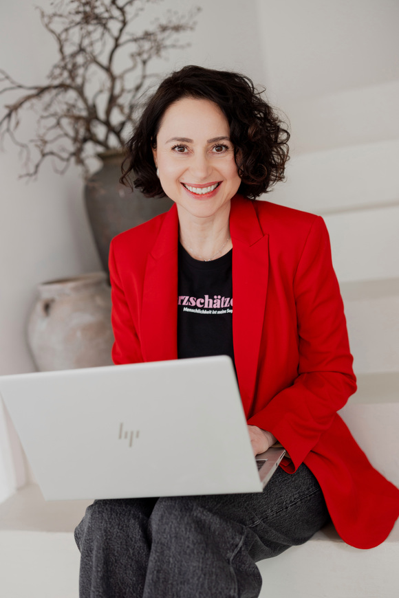 1. Claudia Di Chio, die Herzschätzerin im roten Blazer, auf weißen Stufen sitzend mit Laptop auf den Beinen, lächelnd in die Kamera