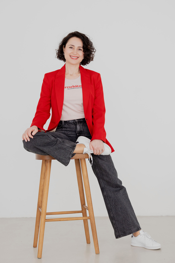 Herzschätzerin, Claudia Di Chio, im roten Blazer, sitzend auf einem Hocker, ein Bein geknickt, eines ausgestreckt