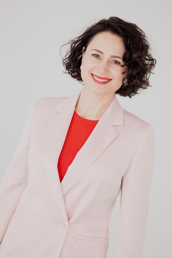 Porträt von Claudia Di Chio, die Herzschätzerin, vor hellem Hintergrund. Sie lächelt in die Kamera und trägt einen hellrosa Blazer über einem roten Oberteil. Ihre dunklen, lockigen Haare reichen bis zum Kinn.